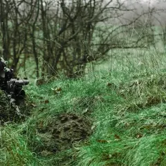 Jugador de airsoft que dispara desde la hierba alta, camuflado y con equipo táctico.